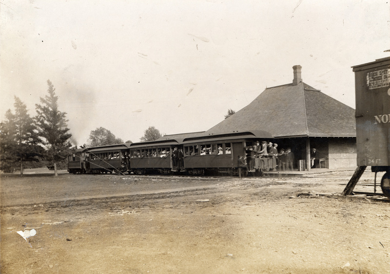 Photograph of the Dinkey stopped in front of the Hub. The engine pulls three passenger cars loaded with students either just arriving on campus or preparing to leave for downtown Ames.