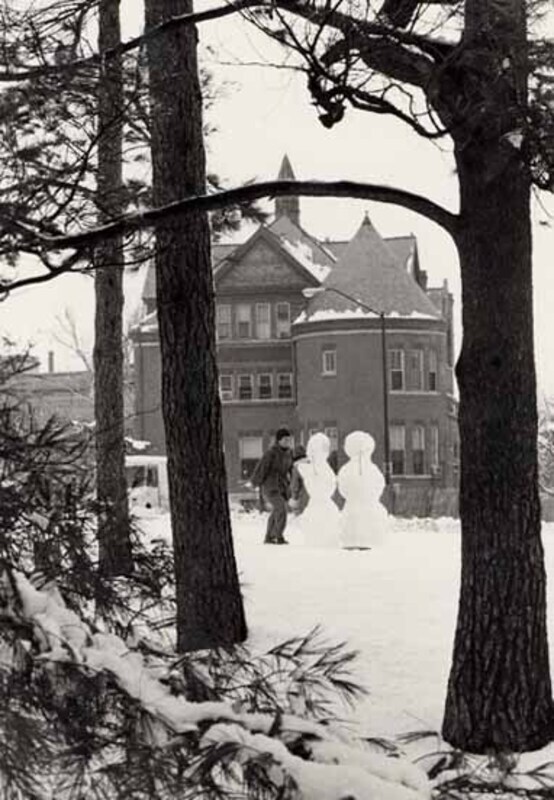 A child in winter clothes stands in front of two snowmen with Morrill Hall in the background.