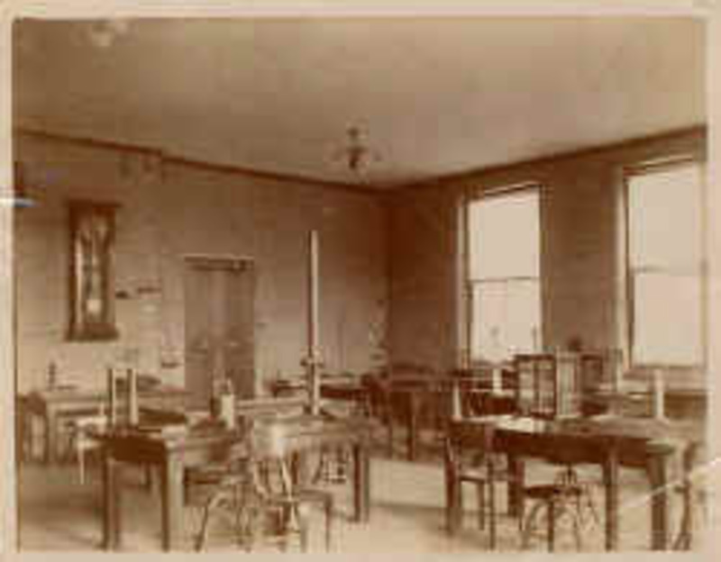 A classroom or lab containing tables, chairs, stools, glass cases, and a wood burning stove.