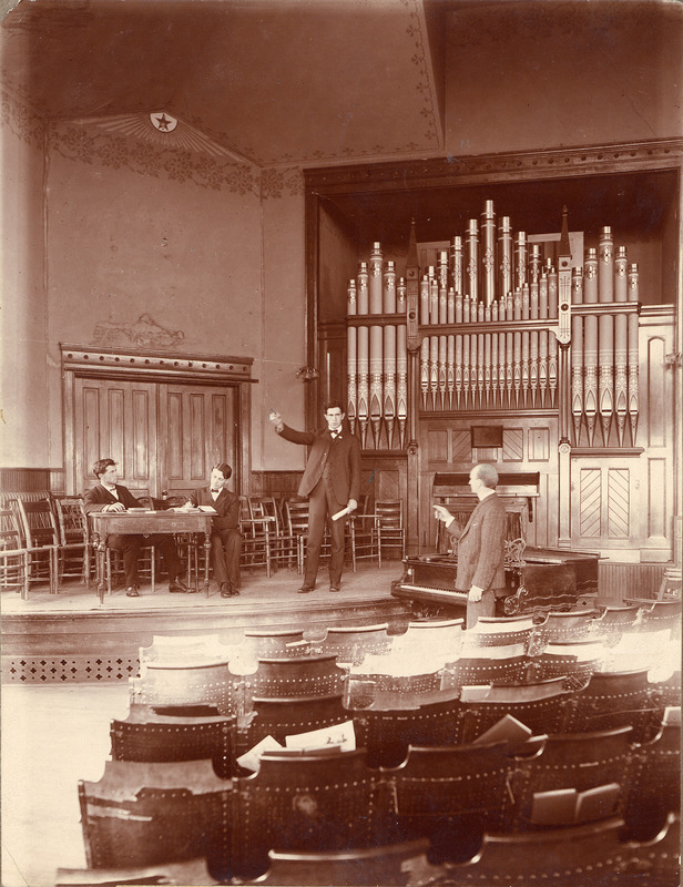 Two men sit at a table onstage in the Morrill Hall chapel in front of the pipe organ. Another man on the stage holds his hand up and gives an oration. A fourth man stands offstage next to a piano.