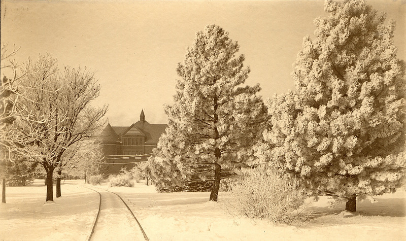 View of the front (east) side of Morrill Hall in winter, partially obscured by trees covered in snow. The tracks of the Ames and College Railway (the Dinkey) are seen leading towards the building.