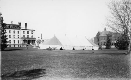A large tent is pictured in the center of this photograph. Old Main is to the left of the tent and Morrill Hall is on the right. Marston Hall (under construction) can be seen in the distance. A man in a horse-drawn wagon is in front of the tent.